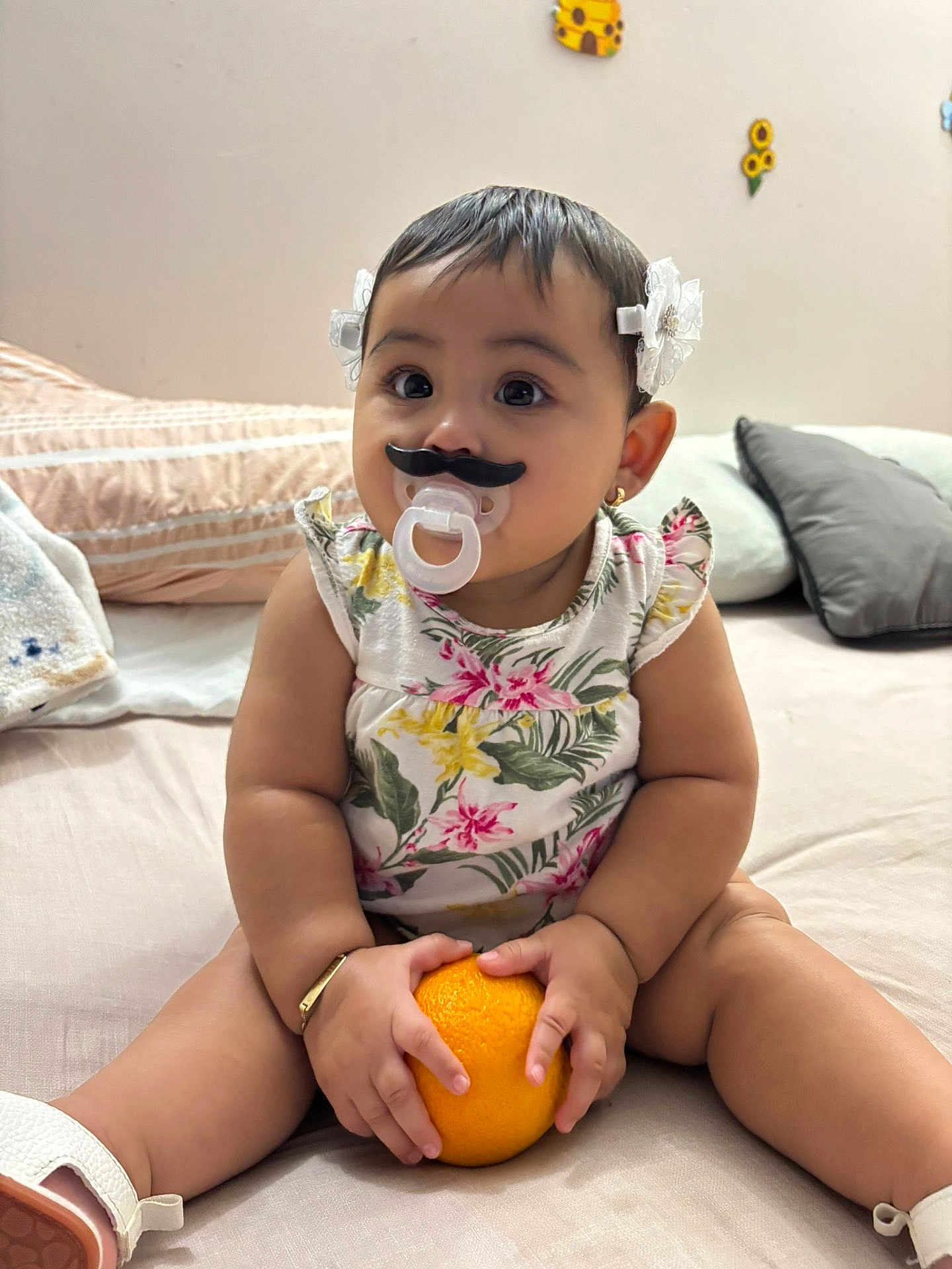 Gianella García is registered to the contest to win money with this photo: baby, bed, bracelet, child, chubby, cute, decor, flower_pattern, hair_bows, indoor, infant, mustache, orange, pacifier, pillow, playful, sitting, smile, toddler, white_shoes
