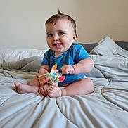 Grayson is registered to the contest to win money with this photo: baby, blue_shirt, toy, bed, pillow, smile, tongue_out, sitting, indoor, child, cute, happy, soft, blanket, person, infant, playful, light_skin, short_hair, room