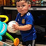 Liam is registered to the contest to win money with this photo: child, toddler, boy, soccer_ball, toy, indoor, standing, holding_ball, shorts, socks, sneakers, sportswear, flowers, vase, table, toy_car, living_room, portrait, curly_hair, cute
