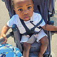 Elijah a rejoint le concours — aidez-le/la à gagner de superbes lots ! child, toddler, stroller, seatbelt, outdoor, sunlight, bag, blue, clothing, shorts, white_shirt, hand, foot, face, eyes, hair, expression, pavement, safety, person