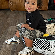 Santos is registered to the contest to win money with this photo: toddler, child, curly_hair, sitting, black_shirt, camouflage_shorts, white_sneakers, nike, cushion, wooden_floor, leather_couch, indoor, cute, smiling, looking_at_camera, floor, patterned_cushion, casual_clothing, young_child, portrait