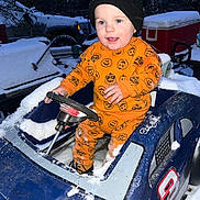 Remington is registered to the contest to win money with this photo: child, toddler, toy_car, snow, winter, orange_clothing, hat, beanie, pumpkin_pattern, outdoor, cold_weather, play, blue_toy, vehicle, happy, smiling, person, snow_covered, steering_wheel, curious