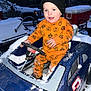 child, toddler, toy_car, snow, winter, orange_clothing, hat, beanie, pumpkin_pattern, outdoor, cold_weather, play, blue_toy, vehicle, happy, smiling, person, snow_covered, steering_wheel, curious