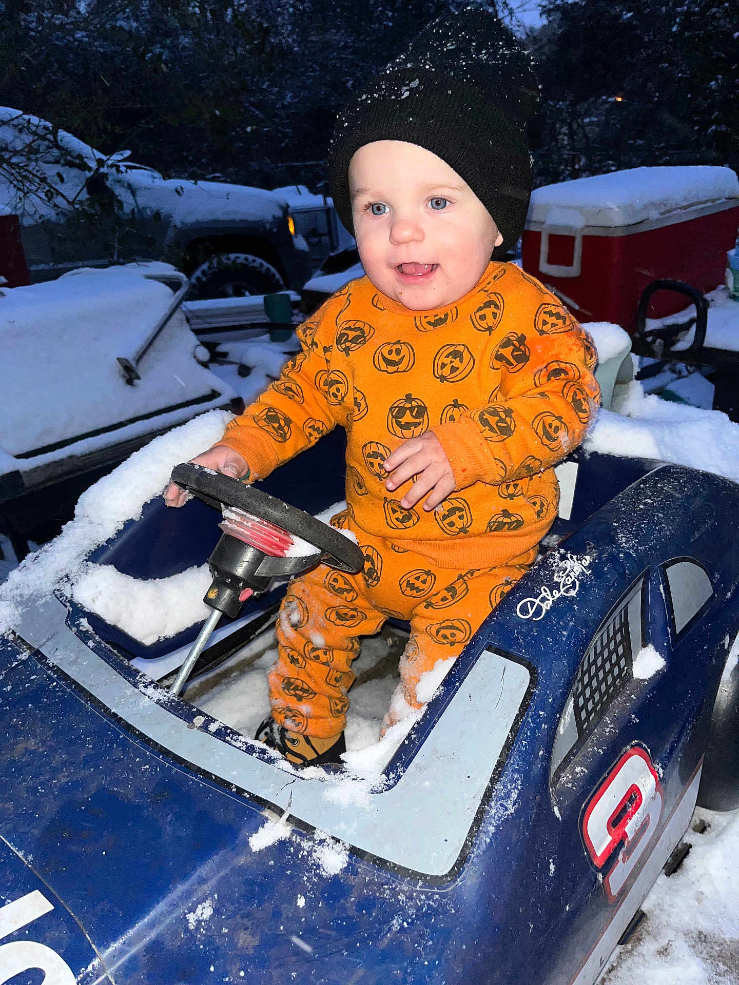 Remington is registered to the contest to win money with this photo: child, toddler, toy_car, snow, winter, orange_clothing, hat, beanie, pumpkin_pattern, outdoor, cold_weather, play, blue_toy, vehicle, happy, smiling, person, snow_covered, steering_wheel, curious