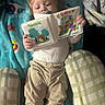 Camden is registered to the contest to win money with this photo: baby, infant, child, reading, book, storybook, shrek, counting, sitting, indoors, blanket, cushion, clothing, pants, white_shirt, face, hands, portrait, cozy, family