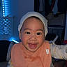 Antonio is registered to the contest to win money with this photo: baby, smiling, bib, cap, indoor, child, happy, face, person, clothing, blue_light, bedroom, furniture, infant, cute, portrait, expression, young_child, reaching, warm_clothing
