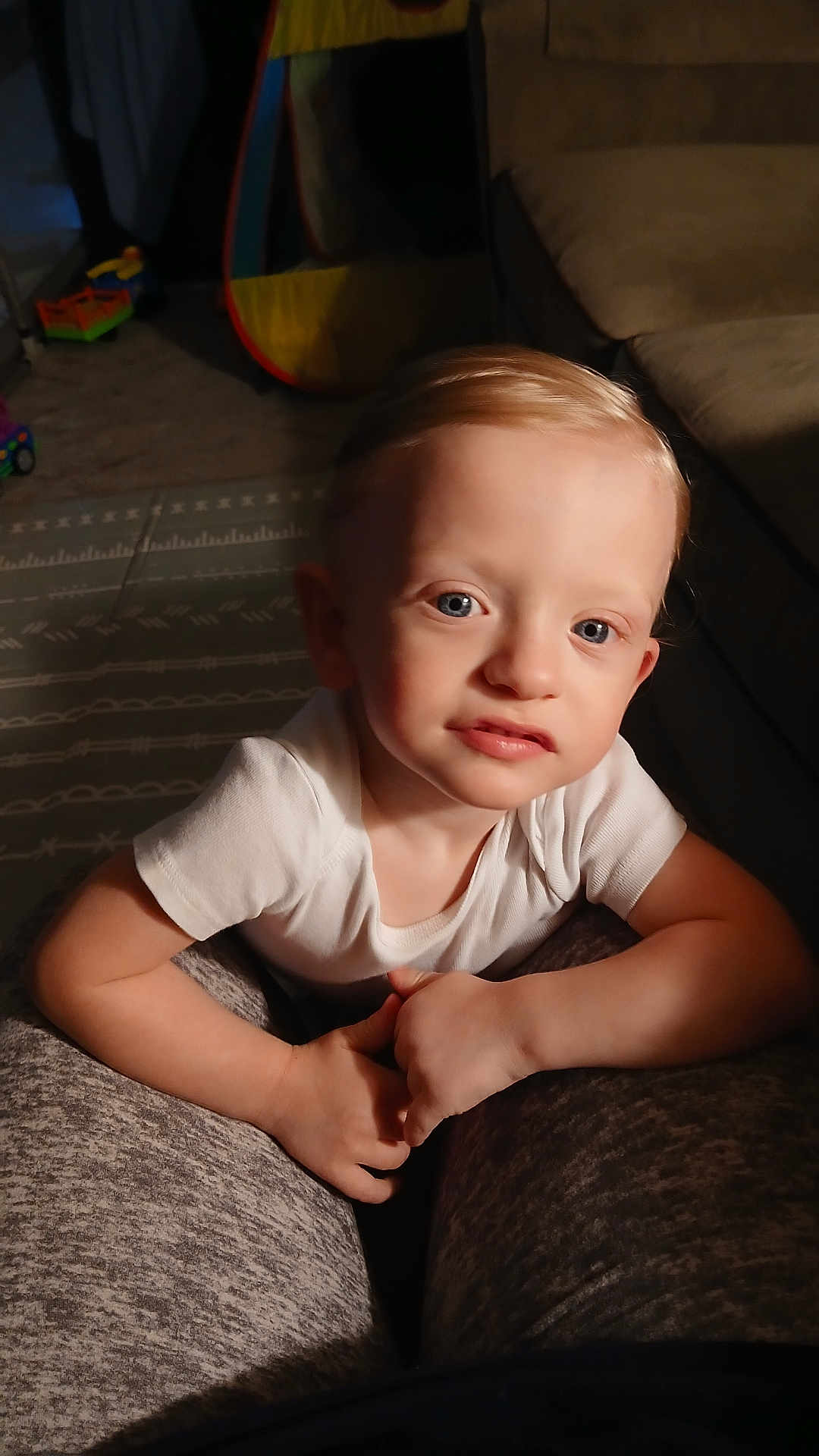 Joseph is registered to the contest to win money with this photo: child, toddler, indoor, person, white_shirt, blonde_hair, blue_eyes, carpet, couch, toy, face, hands, legs, sitting, looking, home, young_child, curious, casual, portrait