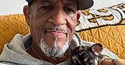 Mickey is registered to the contest to win money with this photo: man, puppy, dog, hoodie, cap, beard, mustache, hand, couch, quilted_cushion, wall, framed_photos, portrait, indoor, smile, pet, casual_clothing, closeup, relaxed, home