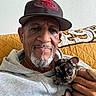 man, puppy, dog, hoodie, cap, beard, mustache, hand, couch, quilted_cushion, wall, framed_photos, portrait, indoor, smile, pet, casual_clothing, closeup, relaxed, home