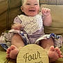 baby, brown_couch, celebration, child, couch, cute, feet, four_months, hands, happy, indoor, infant, lace, milestone, person, portrait, purple_clothing, sitting, smiling, wooden_sign