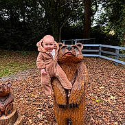 Jesse joined the competition — help win amazing prizes! baby, child, wooden_statue, bear, onesie, forest, autumn, leaves, outdoor, nature, fence, tree, playful, smiling, cute, park, sculpture, person, wood_chips, daytime
