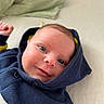 baby, infant, face, smile, hoodie, blue_clothing, lying_down, pillow, closeup, skin, eyes, cheeks, newborn, child, cute, expression, indoors, fabric, soft, portrait