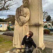 Raphaël participe au concours pour gagner de l'argent avec cette photo : child, statue, stone, outdoor, cloudy_sky, tree, road, car, building, grass, shrub, person, clothing, jacket, pants, footwear, sculpture, monument, curious, daytime