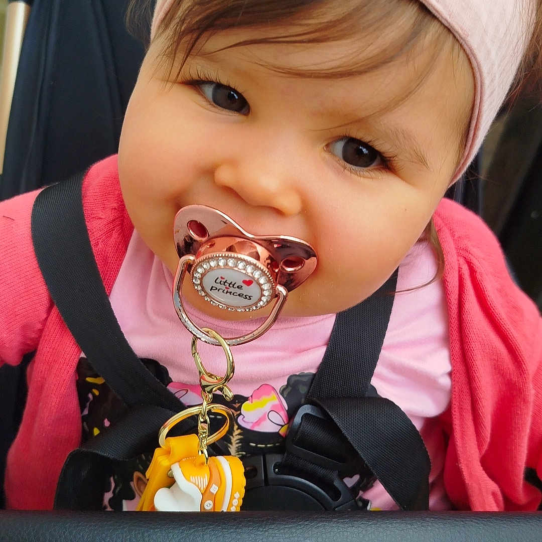 Zheïna participe au concours pour gagner de l'argent avec cette photo : accessory, baby, child, closeup, cute, face, happy, headband, infant, outdoor, pacifier, person, pink_clothing, portrait, seatbelt, smiling, stroller, toddler, toy, young_child