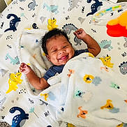 Ja'Rius is registered to the contest to win money with this photo: baby, smiling, blanket, dinosaur, animal_print, infant, lying_down, happy, cozy, cute, child, patterned_sheet, colorful, indoors, playful, soft, warm, toddler, face, raised_arms