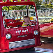 Josiah Pinsonneault is registered to the contest to win money with this photo: child, amusement_park, firetruck, ride, red, smiling, daylight, outdoor, playful, fence, trees, yellow_shirt, young, vehicle, fun, happy, people, park, summer, kid