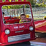child, amusement_park, firetruck, ride, red, smiling, daylight, outdoor, playful, fence, trees, yellow_shirt, young, vehicle, fun, happy, people, park, summer, kid