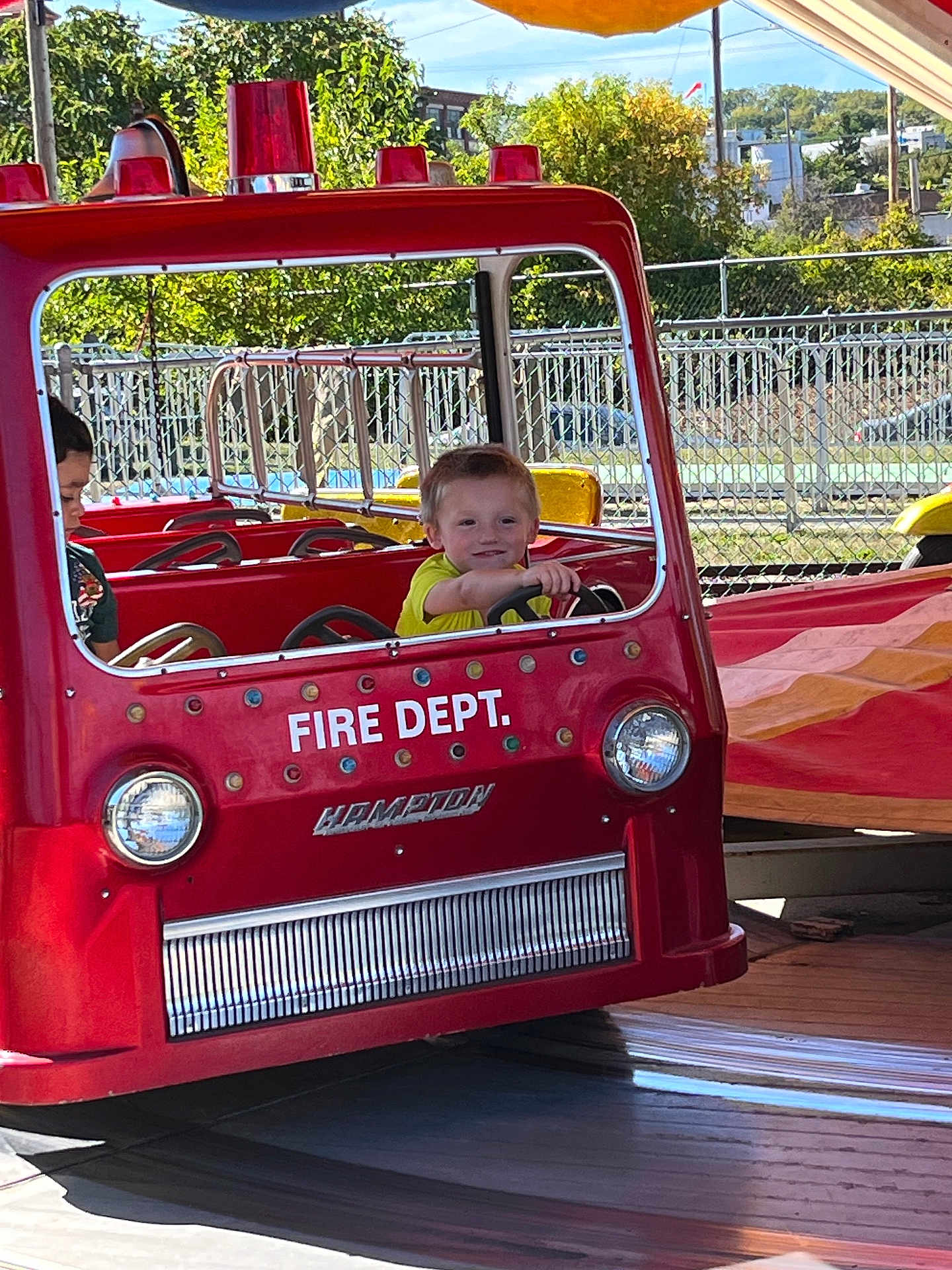 Josiah Pinsonneault is registered to the contest to win money with this photo: child, amusement_park, firetruck, ride, red, smiling, daylight, outdoor, playful, fence, trees, yellow_shirt, young, vehicle, fun, happy, people, park, summer, kid