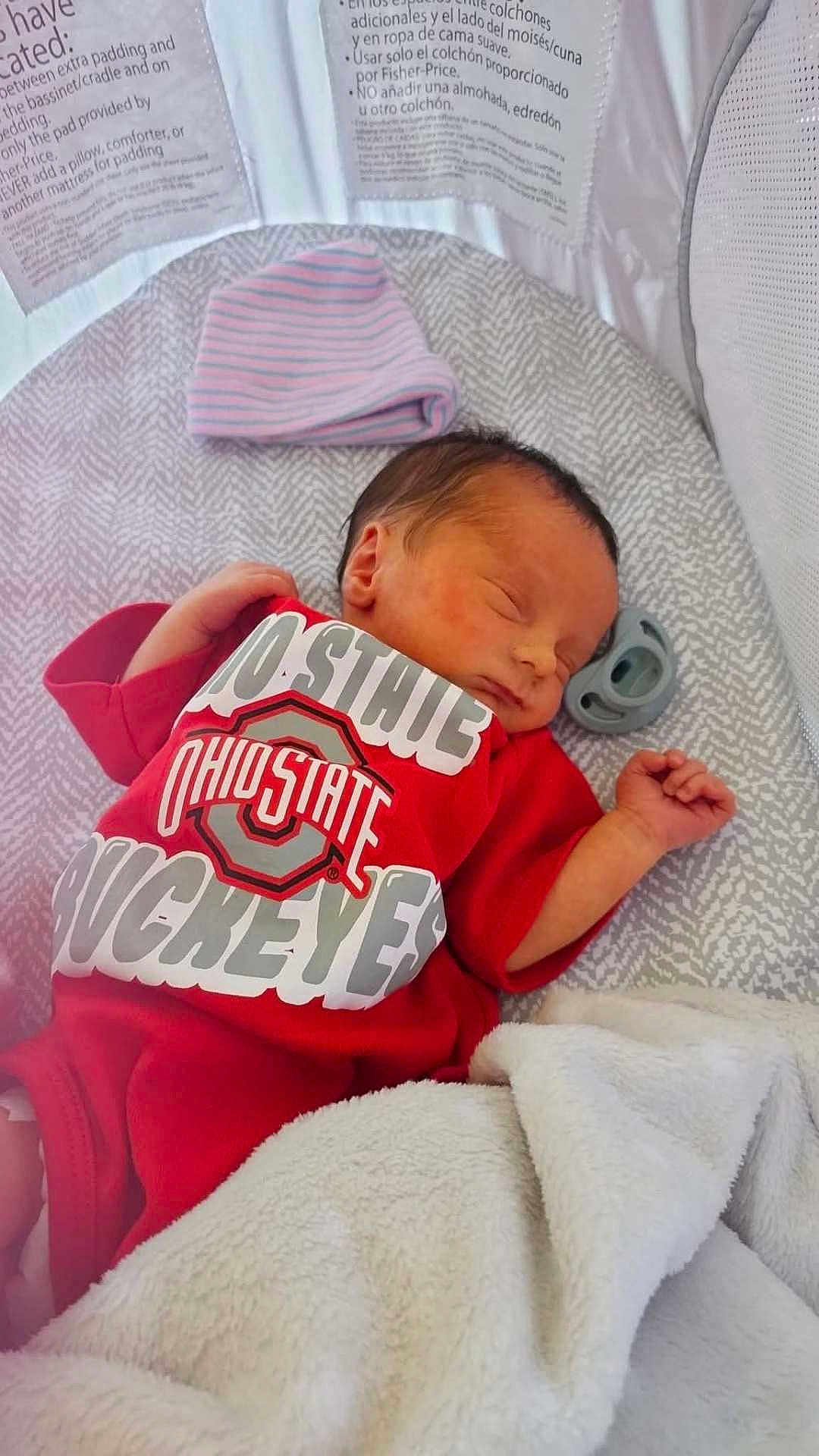 Anthony Allen Pucci is registered to the contest to win money with this photo: newborn, baby, sleeping, onesie, red_clothing, crib, blanket, hat, pacifier, infant, soft_texture, cozy, indoor, resting, cute, peaceful, close_up, face, hand, baby_bedding