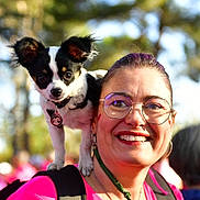 Auguste participe au concours pour gagner de l'argent avec cette photo : woman, dog, glasses, smile, outdoor, shoulder, black_and_white_dog, pink_shirt, sunlight, backpack, happy, person, pet, nature, trees, daylight, portrait, small_dog, people, event