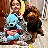 child, dog, birthday_hat, plush_toy, pajamas, birthday_treat, bone_shaped_cookie, indoor, floor, smiling, celebration, pet, toy, happy, cute, sitting, party, home, clothing, fur