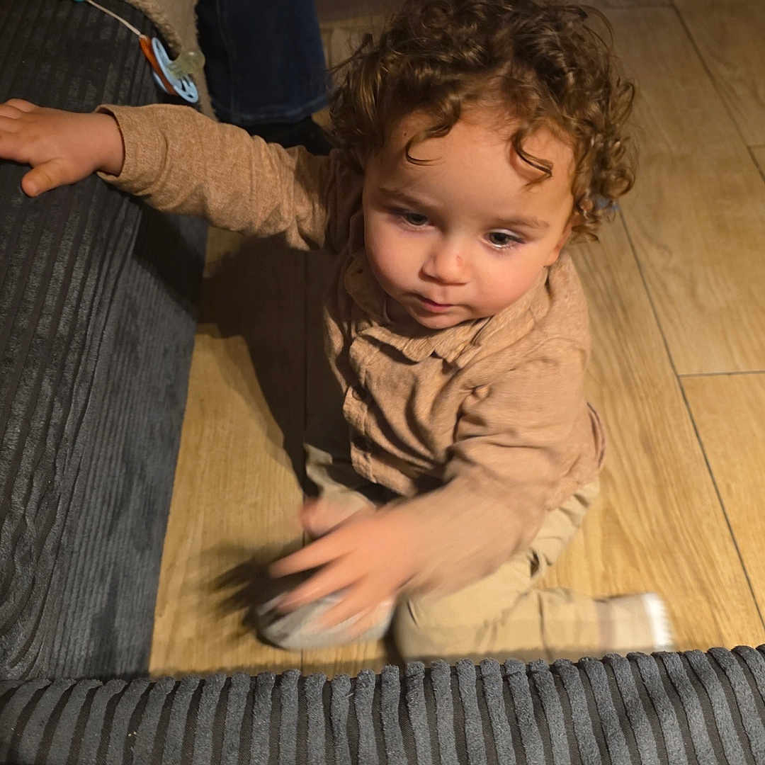 Yassine a rejoint le concours — aidez-le/la à gagner de superbes lots ! adult_leg, child, clothing, couch, curly_hair, face, floor, hand, home, indoor, motion_blur, pacifier, reaching, sitting, slipper, sock, sofa, toddler, toy, wooden_floor