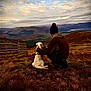 dog, person, mountains, sky, clouds, grass, outdoor, scenery, nature, hill, fence, beanie, jacket, pet, companionship, landscape, rock, sitting, back_view, autumn