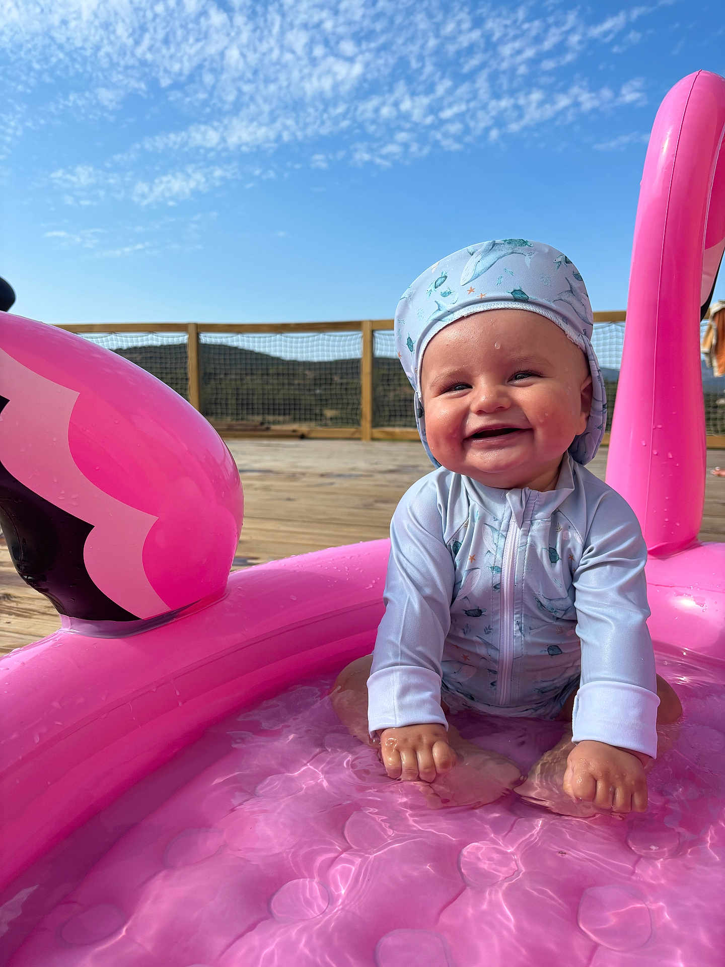 Izak a rejoint le concours — aidez-le/la à gagner de superbes lots ! baby, infant, child, smiling, pink_inflatable, flamingo_pool, water, outdoor, sunny, blue_sky, clouds, wooden_deck, hat, swimwear, happy, sitting, playful, summer, nature, daylight