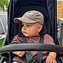baby, child, stroller, cap, harness, outdoor, greenery, wooden_fence, curious, casual_clothing, brown_shirt, shorts, baby_hand, seat, baby_face, summer, daylight, park, baby_boy, infant