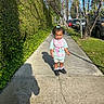 bib, black_shoes, cars, child, curly_hair, daytime, greenery, hedge, nature, outdoor, shadow, shirt, shorts, sidewalk, smiling, street, sunlight, toddler, trees, urban