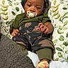 baby, blanket, camo_pants, child, clothing, cozy, cute, face, green_shirt, headwear, indoor, infant, leafy_pillow, lying_down, pacifier, person, portrait, relaxed, soft_texture, young