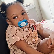 Danny joined the competition — help win amazing prizes! baby, child, infant, portrait, sitting, pacifier, hair_puffs, smiling, indoors, bedroom, bed, clothing, patterned_outfit, blue_pacifier, person, face, eyes, arms, family, home