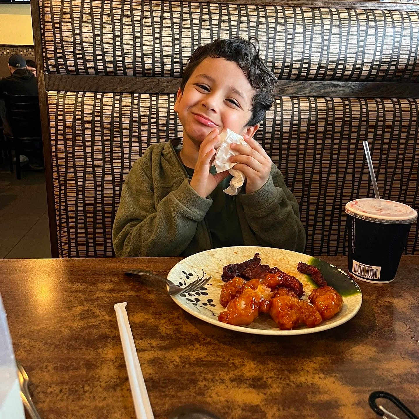 Josh is registered to the contest to win money with this photo: child, smiling, restaurant, booth, food, plate, drink, straw, napkin, utensils, table, dining, indoor, portrait, curly_hair, green_sweatshirt, soccer, television, happy, family_meal