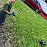 child, boy, outdoor, backyard, grass, lawn, toy, remote_control, toy_car, rc_truck, vehicle, red_car, house, driveway, greenery, standing, playing, casual_clothing, sneakers, portrait