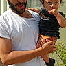child, toddler, adult, man, father, parent, portrait, outdoor, sunlight, family, holding, standing, smiling, beard, black_shirt, white_shirt, window, house, potted_plant, photography
