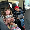 children, child, car, back_seat, smiling, happy, siblings, winter_clothing, red_hat, beanie, tablet, doll, toy, seat_belts, vehicle, road_trip, luggage, portrait, indoors, family