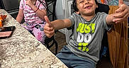 Josh is registered to the contest to win money with this photo: children, boy, girl, smiling, thumbs_up, portrait, indoor, kitchen, table, countertop, refrigerator, groceries, shelves, chair, gray_shirt, pink_pajamas, curly_hair, long_hair, happy, family