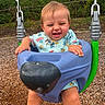 baby, child, infant, smiling, laughing, playground, swing, bucket_swing, outdoor, park, playground_equipment, swing_chains, wood_chips, greenery, trees, blue_clothing, patterned_shirt, seated, portrait, happy