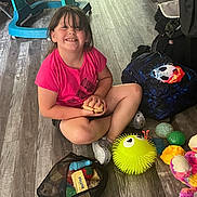 Olivia joined the competition — help win amazing prizes! child, smiling, indoors, floor, toys, backpack, soccer_ball, snack, pink_shirt, seated, playroom, wood_floor, toy_ball, stuffed_animal, spiky_toy, baby_walker, household, portrait, happy, playtime
