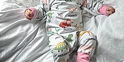 Evelyn joined the competition — help win amazing prizes! baby, bedroom, child, cozy, cute, dinosaur_pajamas, happy, indoors, infant, infant_clothing, joyful, lying_down, mouth_open, one_person, pajamas, playful, small_feet, smiling, soft_lighting, white_bed