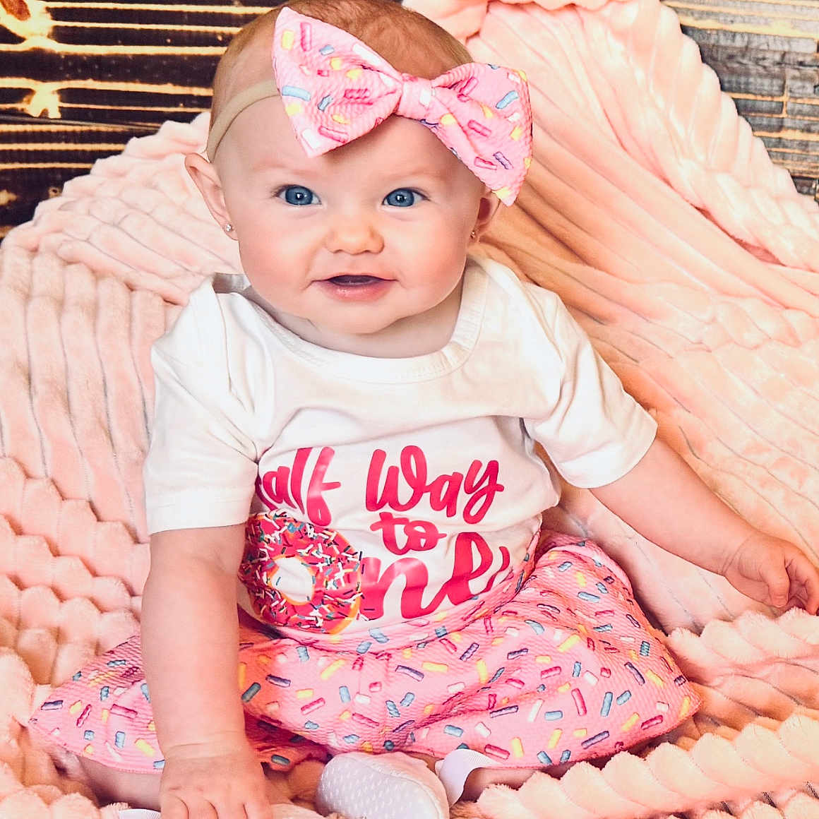 Lilyanne is registered to the contest to win money with this photo: baby, blanket, blue_eyes, child, colorful, cute, donut_graphic, footwear, happy, headband, indoor, infant, pink_bow, pink_skirt, portrait, sitting, smiling, soft_texture, white_shirt, wood_background