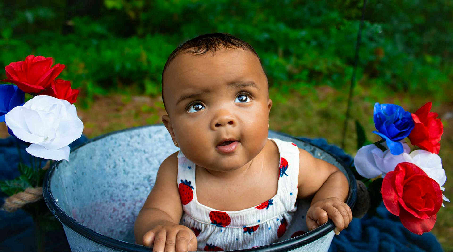 Ta'Maya joined the competition — help win amazing prizes! baby, child, bucket, flowers, red_flower, white_flower, blue_flower, outdoor, greenery, garden, curious, infant, portrait, summer_clothing, strawberry_pattern, hands, face, skin, nature, cute