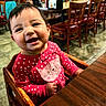 baby, child, smiling, high_chair, pink_clothing, polka_dot, cat_design, wooden_chair, restaurant, table, condiments, happy, cute, indoor, floor_tiles, person, face, seated, dining_area, bright