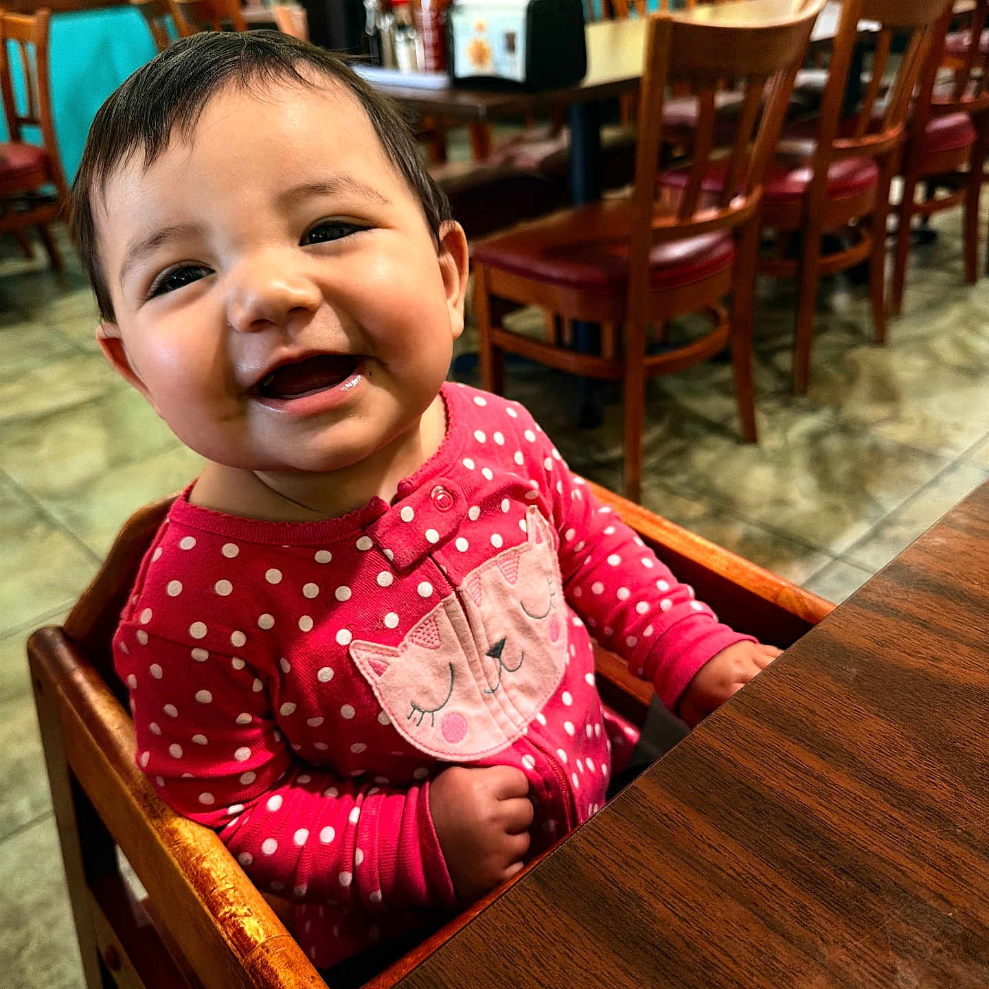 Asyla is registered to the contest to win money with this photo: baby, child, smiling, high_chair, pink_clothing, polka_dot, cat_design, wooden_chair, restaurant, table, condiments, happy, cute, indoor, floor_tiles, person, face, seated, dining_area, bright