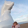 child, sculpture, white, animal_statue, beach, sand, sky, clouds, jacket, hugging, outdoor, art, statue, winter_clothing, cute, small_child, daytime, expression, playful, landscape