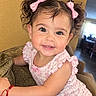 child, toddler, girl, smiling, portrait, indoors, couch, sitting, pink_bows, floral_dress, curly_hair, brown_hair, eyes, happy, baby, home, furniture, close_up, photograph, phone_screenshot