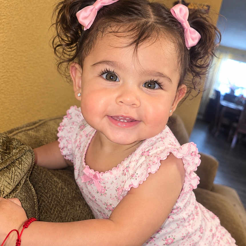Celina is registered to the contest to win money with this photo: child, toddler, girl, smiling, portrait, indoors, couch, sitting, pink_bows, floral_dress, curly_hair, brown_hair, eyes, happy, baby, home, furniture, close_up, photograph, phone_screenshot