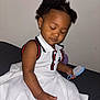 toddler, child, dress, white_dress, hairbrush, sitting, closed_eyes, curly_hair, earrings, indoor, calm, portrait, person, fashion, black_hair, brown_skin, furniture, bed, relaxed, cute