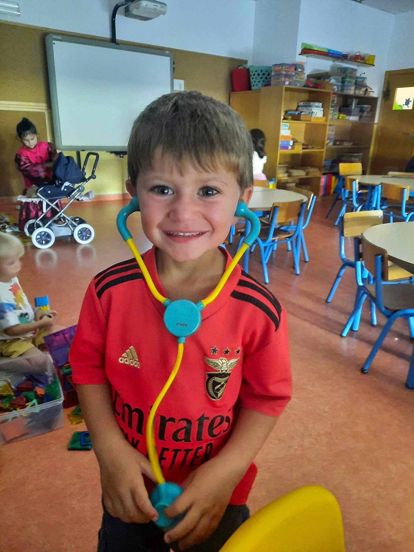 Dani participe au concours pour gagner de l'argent avec cette photo : child, boy, smiling, toy, stethoscope, classroom, school, play, red_shirt, furniture, chairs, tables, toys, kids, indoor, education, happy, person, casual_clothing, learning