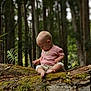 Mavis is registered to the contest to win money with this photo: baby, barefoot, casual_clothing, child, curious, daylight, exploring, forest, greenery, leaves, log, moss, nature, outdoor, pink_shirt, sitting, tree, trunk, woodland, young_child